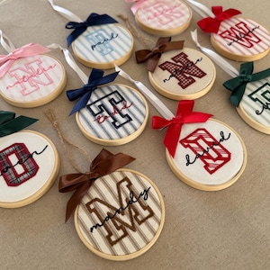 Könnte beinhalten: Eine Sammlung runder Stickrahmen-Ornamente aus Holz mit Stoffeinsätzen, die mit gestickten Blockbuchstaben und kursiven Namen versehen sind. Jedes Ornament ist mit einer Satinbandschleife in verschiedenen Farben wie Rot, Marineblau, Braun, Rosa und Dunkelgrün verziert.