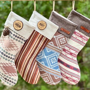 May include: Four Christmas stockings with personalized wooden name tags and various patterned designs. The stockings are in shades of brown, red, and blue, with a neutral linen cuff. One stocking has a pinecone attached. Each stocking is hanging from a wooden rod.