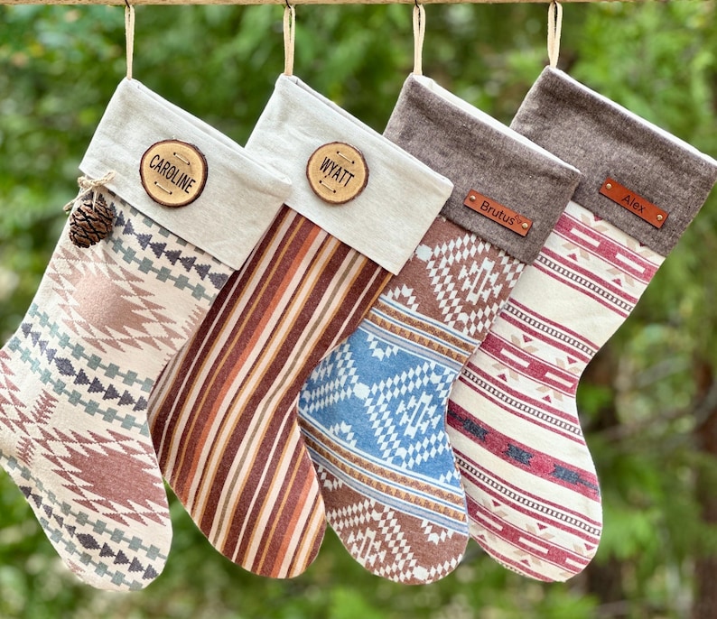 May include: Four hanging Christmas stockings with varied patterns. Two have cream cuffs with wooden name tags reading Caroline and Wyatt. Two have brown cuffs with leather tags reading Brutus and Alex. The stockings feature geometric and striped designs.