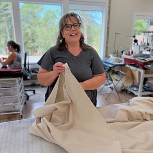 May include: A person wearing a dark gray v-neck t-shirt and glasses, holding a large piece of solid beige fabric. The fabric is draped over a cutting table with a grid pattern, showcasing its smooth texture and neutral color in a bright workspace.