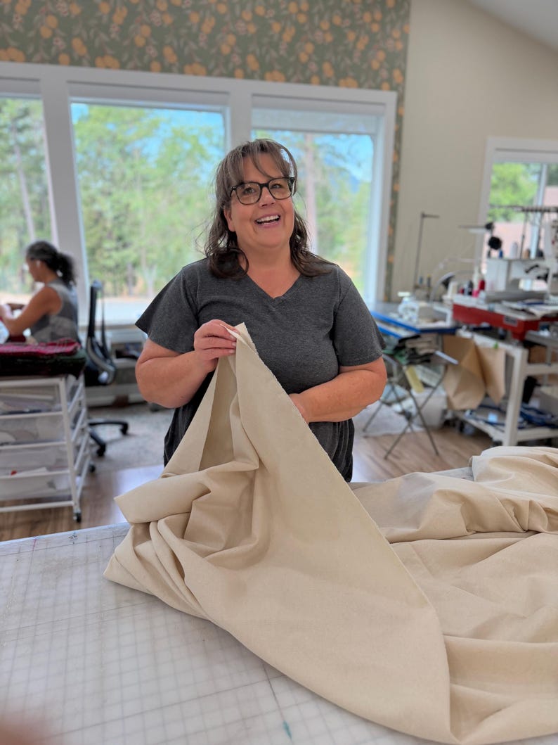 May include: A person wearing a dark gray v-neck t-shirt and glasses, holding a large piece of solid beige fabric. The fabric is draped over a cutting table with a grid pattern, showcasing its smooth texture and neutral color in a bright workspace.