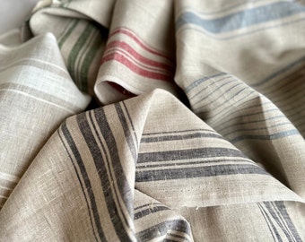 May include: A collection of folded linen fabric pieces in a natural beige tone, each featuring distinct vertical stripes in various colors including red, blue, green, and charcoal gray. The fabric has a visible woven texture and soft folds.