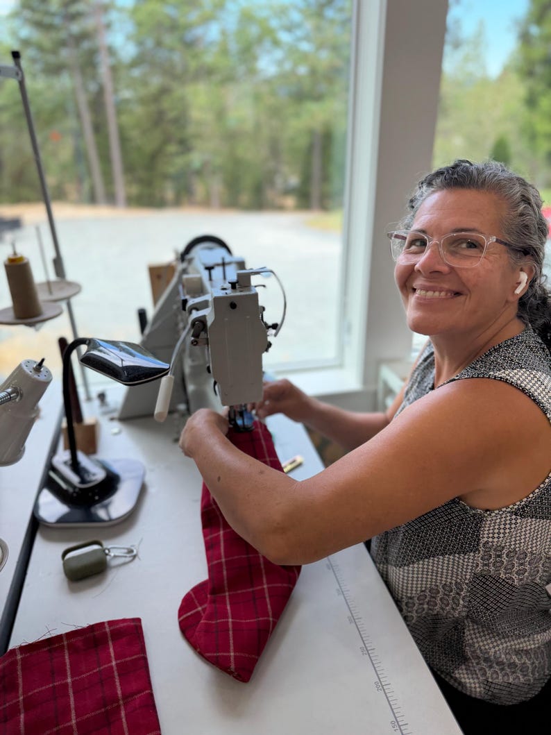 May include: A grey industrial sewing machine with a black task lamp and a spool of thread on a white table. A piece of red plaid fabric is being fed through the machine needle. A small olive green case with a silver carabiner sits on the table.