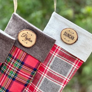 May include: Two Christmas stockings featuring red, green, and white plaid fabric with solid-colored cuffs. One stocking has a dark brown cuff with a wooden slice tag labeled Peyton, and the other has a light gray cuff with a wooden tag labeled DUNCAN.