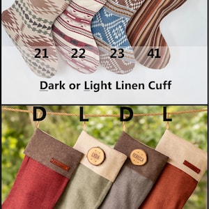 K&ouml;nnte beinhalten: Acht Weihnachtsstr&uuml;mpfe mit verschiedenen Mustern und Unifarben, ausgestattet mit dunklen oder hellen Leinenb&uuml;ndchen. Einige Str&uuml;mpfe haben personalisierte Namensschilder aus Holz oder Leder. Die Str&uuml;mpfe sind mit 21, 22, 23, 41, 25, 26, 27 und 28 nummeriert.