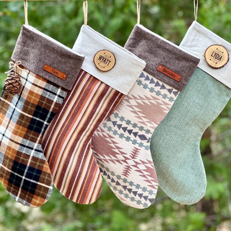 K&ouml;nnte beinhalten: Vier h&auml;ngende Weihnachtsstr&uuml;mpfe mit personalisierten Anh&auml;ngern. Der erste ist kariert mit braunem Umschlag und einem Tannenzapfen. Der zweite hat vertikale Streifen und einen hellen Umschlag mit einem Holzanh&auml;nger mit der Aufschrift WYATT. Der dritte hat ein geometrisches Muster und einen braunen Umschlag mit einem Anh&auml;nger mit der Aufschrift Brooks. Der vierte ist einfarbig salbeigr&uuml;n mit hellem Umschlag und einem Holzanh&auml;nger mit der Aufschrift LYDIA.