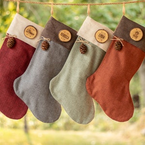 May include: Four personalized Christmas stockings hanging on a rope. Each stocking is a different color: burgundy, gray, sage green, and rust. They have a cream-colored cuff and a wooden name tag. Each stocking is decorated with a small pinecone.