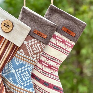 May include: Three Christmas stockings featuring varied patterns and personalized tags. One has a wood slice tag labeled WYATT, while the others have leather-like tags labeled Brutus and Alex. The stockings display striped and geometric designs.