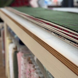 K&ouml;nnte beinhalten: Ein Stapel gefalteter Stoffst&uuml;cke in verschiedenen Farben und Mustern, darunter Gr&uuml;n, Rot und Wei&szlig;, auf einem hellen Holztisch. Unter dem Tisch sind mehrere Stoffrollen in Regalen in einem Lagerbereich organisiert.