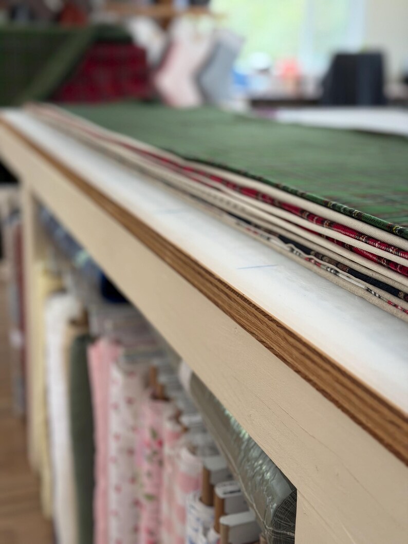 K&ouml;nnte beinhalten: Ein Stapel gefalteter Stoffst&uuml;cke in verschiedenen Farben und Mustern, darunter Gr&uuml;n, Rot und Wei&szlig;, auf einem hellen Holztisch. Unter dem Tisch sind mehrere Stoffrollen in Regalen in einem Lagerbereich organisiert.