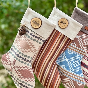 May include: Four personalized Christmas stockings with various patterns and colors. Each stocking has a wooden name tag and is suspended by twine. The stockings feature designs in shades of brown, blue, and cream, with a pinecone accent on one.