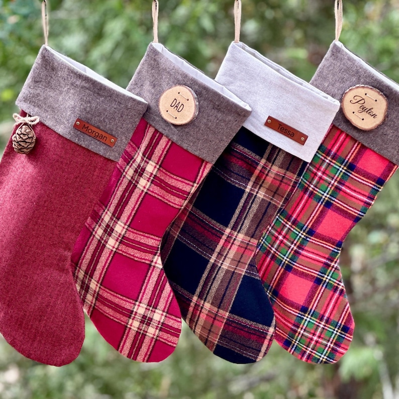 May include: Four Christmas stockings hanging in a row. The first is solid red with a brown cuff and a small pinecone. The second is red plaid with a brown cuff and a wood slice tag labeled DAD. The third is navy plaid with a light cuff and a leather tag labeled Tessa. The fourth is red and green plaid with a brown cuff and a wood slice tag labeled Peyton. The first has a leather tag labeled Morgan.