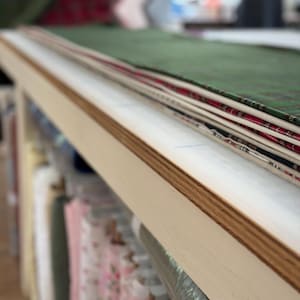 May include: A stack of folded fabric pieces in various colors and patterns, including green, red, and white, resting on a light-colored wooden table. Below the table, multiple rolls of fabric are organized on shelves in a storage area.