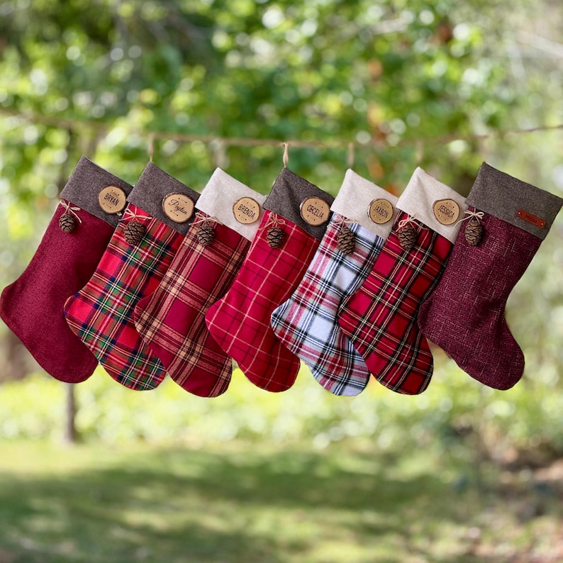 May include: Seven Christmas stockings hanging in a row, featuring various red, white, and green plaid patterns and solid burgundy fabric. Each stocking has a brown cuff, a small pinecone accent, and a round wooden tag engraved with a name.