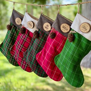 May include: Five hanging Christmas stockings in alternating red and green plaid patterns. Each stocking features a neutral-colored cuff, a small brown pinecone accent, and a round wooden name tag engraved with names including Angel, Jessica, Harley, Cecelia, and Riley.
