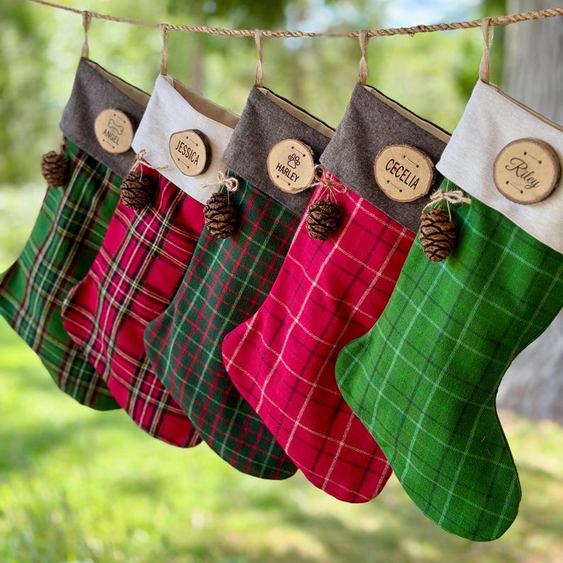 May include: Five hanging Christmas stockings in alternating red and green plaid patterns. Each stocking features a neutral-colored cuff, a small brown pinecone accent, and a round wooden name tag engraved with names including Angel, Jessica, Harley, Cecelia, and Riley.