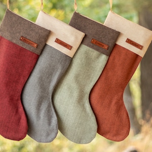 May include: Four hanging Christmas stockings in red, gray, sage green, and burnt orange fabric. Each stocking features a contrasting cuff with a brown leather name tag attached by two metal rivets. The names Morgan, Jonah, Emma, and Caden are printed.
