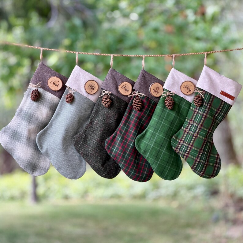 May include: A row of six hanging stockings featuring various plaid and solid fabric patterns in shades of green, gray, and brown. Each stocking is topped with a light-colored cuff, a small wooden name tag, and a decorative brown pinecone accent.