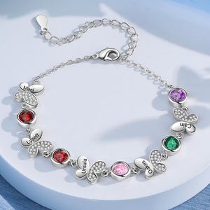 May include: A silver bracelet featuring butterfly-shaped charms with embedded gemstones in red, pink, green, and purple. Each butterfly has a name engraved on it. The bracelet is displayed on a white surface.