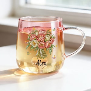 May include: A clear glass mug with a pink-tinted rim, featuring a printed illustration of a bouquet of pink and white peonies with small yellow and purple butterflies. The name Alex is printed in black script on a heart shape below the flowers.