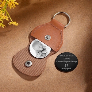 May include: A tan leather keychain with a metal ring and a snap closure. Inside the open leather flap is a circular black and white photo of a baby. Beside it lies a round black metal disc engraved with the text POCKET HUG Daddy I am with you always Baby Jack.