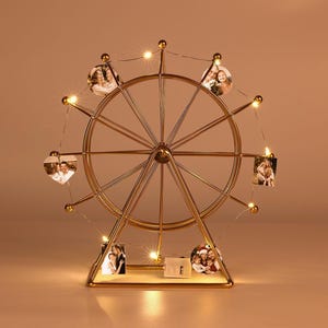 May include: A gold-colored metal ferris wheel photo display stand featuring a circular frame with spokes. The structure is illuminated by thin string lights and includes several small circular and heart-shaped photographs clipped to the outer rim.