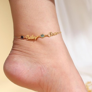 May include: A gold-colored anklet with the names "Bailey" and "Kirsten" inscribed. The anklet features a small, dark red gemstone and a light blue gemstone. The jewelry is worn on an ankle.