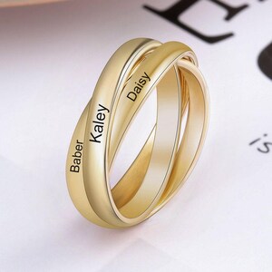 May include: A gold-toned triple interlocking band ring featuring three smooth, polished metal loops. Each band is engraved with a name, specifically the names Baber, Kaley, and Daisy, displayed in a clear, dark, sans-serif font on the outer surface.