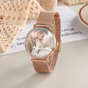 K&ouml;nnte beinhalten: Ros&eacute;goldene Armbanduhr mit einem Mesh-Armband. Das Zifferblatt zeigt ein Foto eines sich k&uuml;ssenden Paares. Die Uhr hat ein rundes Zifferblatt mit einer kristallbesetzten L&uuml;nette und zarten Zeigern.
