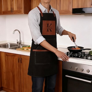 May include: A black apron featuring brown leather straps and trim, with a square brown leather patch on the chest displaying the letter K, the name Kevin, and the letter K again. The apron includes a large front pocket with brown leather accents.