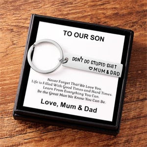 May include: A silver-toned metal keychain with a rectangular tag engraved with the text 'DON'T DO STUPID SHIT' and a heart symbol followed by 'MUM & DAD'. The keychain is presented in a black box with a card reading 'TO OUR SON' and a message.