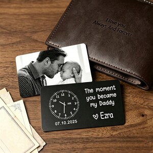 May include: A dark brown leather wallet with the inscription I love you Always And Forever, paired with two rectangular metal cards. One card features a black and white photo of a person and a baby, while the other is black with a clock graphic, the text The moment you became my Daddy, the name Ezra, and the date 07.10.2025.