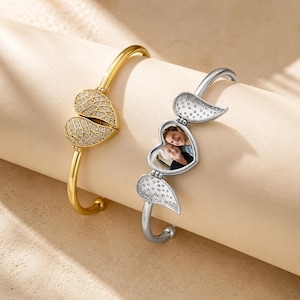 May include: Two bangle bracelets, one gold-toned and one silver-toned, each featuring a heart-shaped locket with angel wing accents encrusted with small clear stones. The silver locket is open, revealing a small photograph of two people inside.