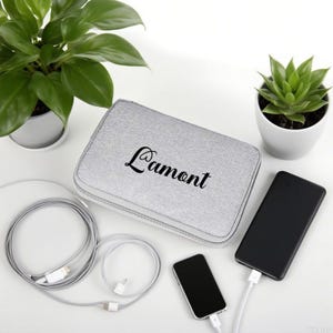 May include: A rectangular light gray fabric storage case with a zipper closure and the name Lamont printed in black script on the top surface. The case is accompanied by a long gray charging cable, a small black smartphone, and a large black power bank.