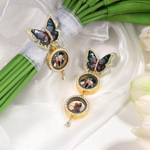May include: Gold-colored butterfly-shaped photo locket with a sparkling rhinestone border. The butterfly has a blue and black wing design. The locket is attached to a bouquet of green stems wrapped in white ribbon. Each locket holds a small photo.