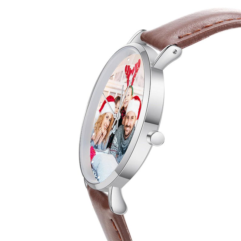 May include: A silver wristwatch with a brown leather band. The watch face displays a family photo. The watch has a round face and a small button on the side. The band is a medium brown color.