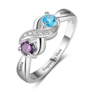 May include: Silver infinity ring with two gemstones, one blue and one purple. The ring is engraved with the names "Susan" and "Anna" and the text "Engraving Here".