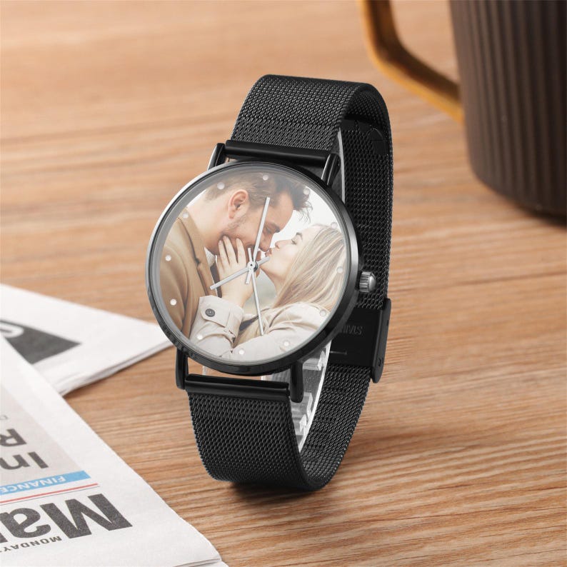 K&ouml;nnte beinhalten: Schwarze Armbanduhr mit einem Mesh-Armband. Das Zifferblatt zeigt ein Farbfoto eines sich k&uuml;ssenden Paares. Die Uhr hat eine schwarze L&uuml;nette und silberne Zeiger. Die Uhr liegt auf einer Holzoberfl&auml;che.