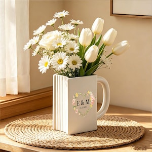 May include: A white ceramic mug-shaped vase with vertical ribbed detailing, featuring a floral heart graphic with the letters E & M and the date 11.22.2025. The vase is filled with a bouquet of white daisies, a white rose, and white tulips.
