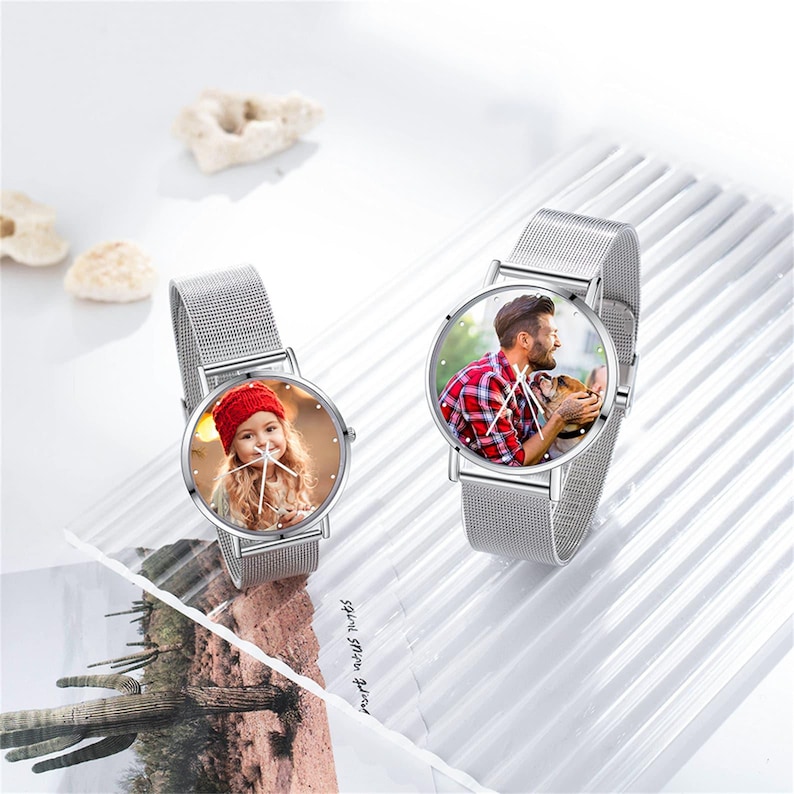 K&ouml;nnte beinhalten: Zwei silberne Armbanduhren mit Mesh-Armb&auml;ndern. Die Zifferbl&auml;tter zeigen personalisierte Fotos: eines von einem jungen M&auml;dchen mit roter M&uuml;tze und das andere von einem Mann mit einem Hund. Die Uhren liegen auf einer wei&szlig;en Oberfl&auml;che.