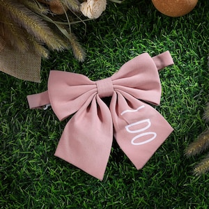 May include: A dusty rose fabric bow tie featuring the white text I DO printed on the right side. The bow tie has a structured, multi-layered design with a central knot and two long, wide tails extending downward, attached to a matching fabric strap.
