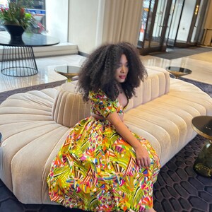 K&ouml;nnte beinhalten: Eine Frau tr&auml;gt ein farbenfrohes, geometrisches Zweiteiler-Outfit mit einem kurz&auml;rmeligen Oberteil und einem Rock. Sie sitzt auf einem beigen Sofa mit einem schwarz-wei&szlig;en gemusterten Teppich.