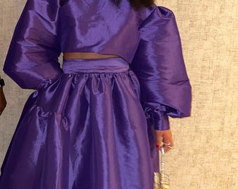 Könnte beinhalten: Ein leuchtend violettes zweiteiliges Outfit mit einem Crop-Top mit voluminösen Ärmeln und einem passenden Midirock. Das Ensemble ist aus einem glänzenden Stoff gefertigt. Das Outfit wird durch weißspitzige Absätze und eine kleine, verzierte Handtasche ergänzt.