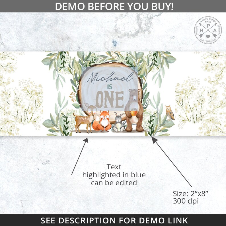 May include: A woodland-themed first birthday banner with a blue and white woodgrain sign that says "Michael is ONE". The sign is surrounded by green leaves and branches, and there are cute animal illustrations including a deer, fox, rabbit, and bear.