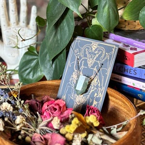 May include: A green crystal pendant necklace with a silver chain. The pendant is shaped like a long, thin rectangle and is set against a black and white illustration of a demon on a card.