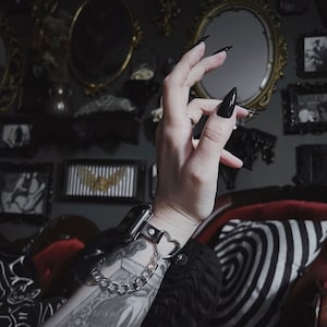 May include: A black leather watch band with a silver chain and a heart-shaped charm. The band is worn on a wrist with a tattoo.