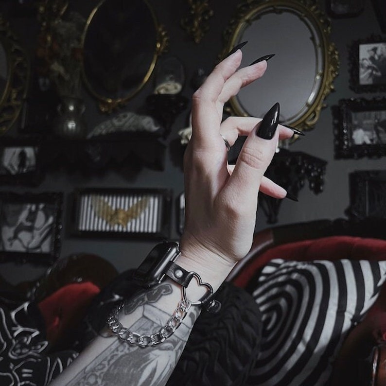 May include: A black leather watch band with a silver chain and a heart-shaped charm. The band is worn on a wrist with a tattoo.
