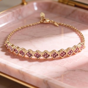 May include: A gold-toned bracelet with a series of pink floral-shaped embellishments. The bracelet is displayed on a pink marble tray with gold trim. The tray is next to a gold-framed mirror.