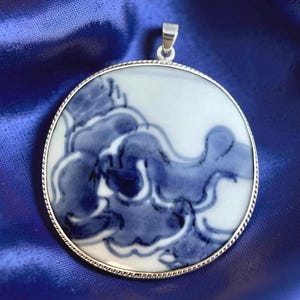 May include: Round pendant with a blue and white floral design. The pendant has a silver-colored metal frame and a bail for a chain. The design features a stylized cloud or wave pattern in shades of blue on a white background.
