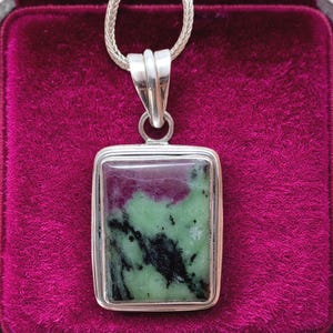 Puede incluir: Colgante rectangular de plata con cadena. La piedra natural presenta colores verdes, negros y rojo-púrpura oscuro. La joya se encuentra sobre un fondo de terciopelo rosa oscuro, resaltando los colores.